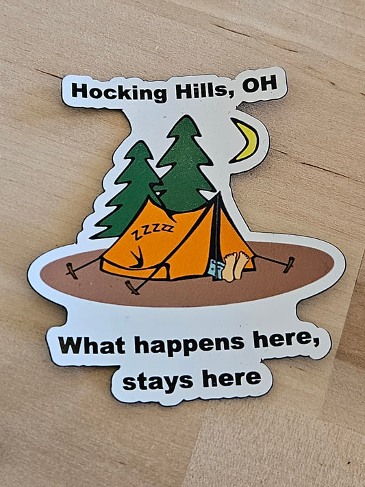 Sticker with a cartoon design of a tree and umbrella, text 'Hocking Hills, OH' and 'What happens here, stays here', on a wooden surface.