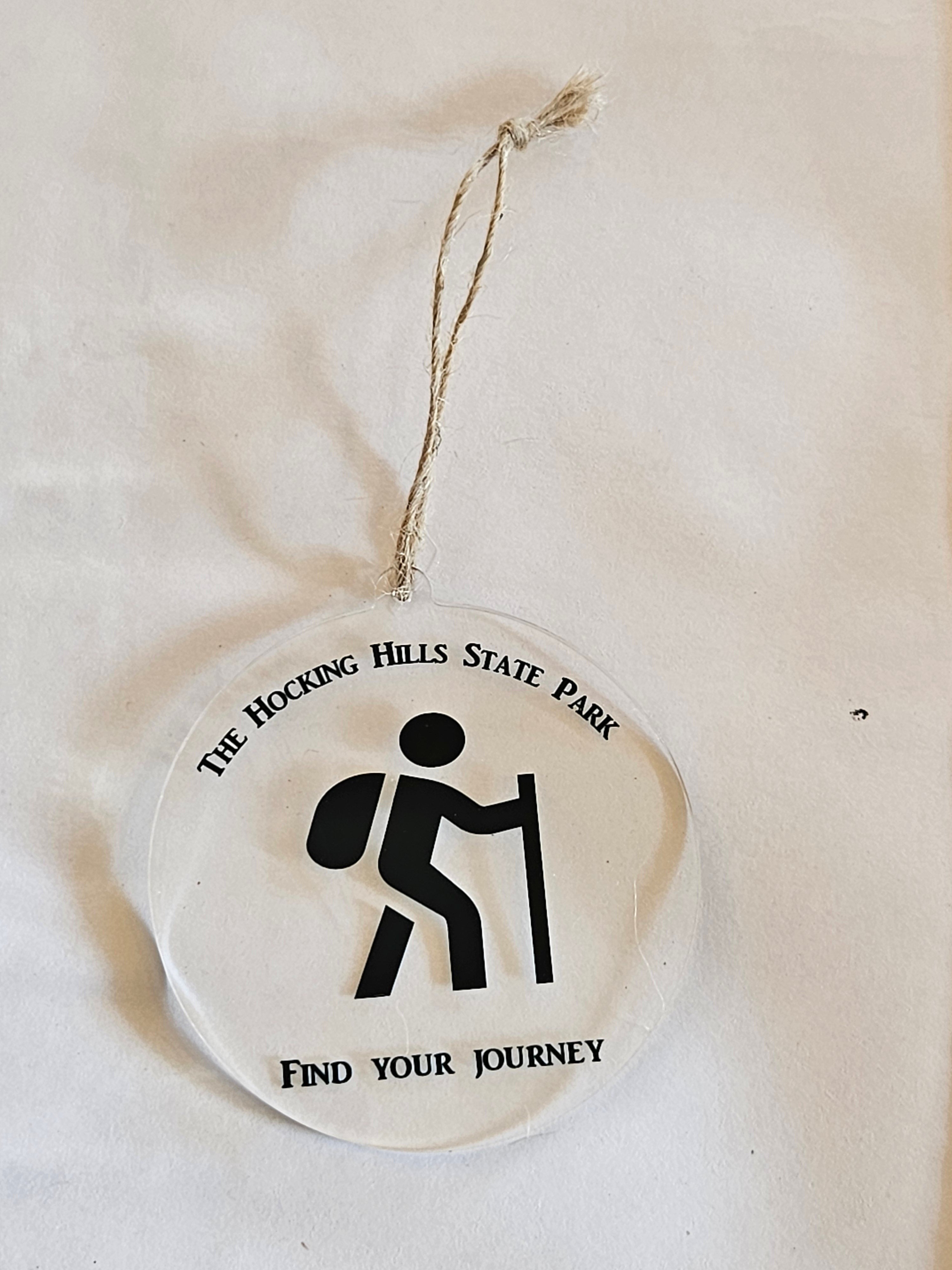 The Hocking Hills Store clear acrylic round ornament with a black hiker icon and text 'THE HOCKING HILLS STATE PARK FIND YOUR JOURNEY'.