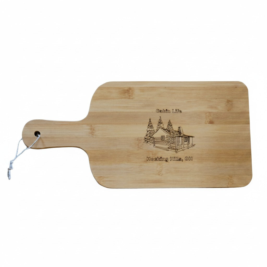 The Hocking Hills Store bamboo cutting board with laser engraved cabin and trees design. Features a handle with a hole for hanging.