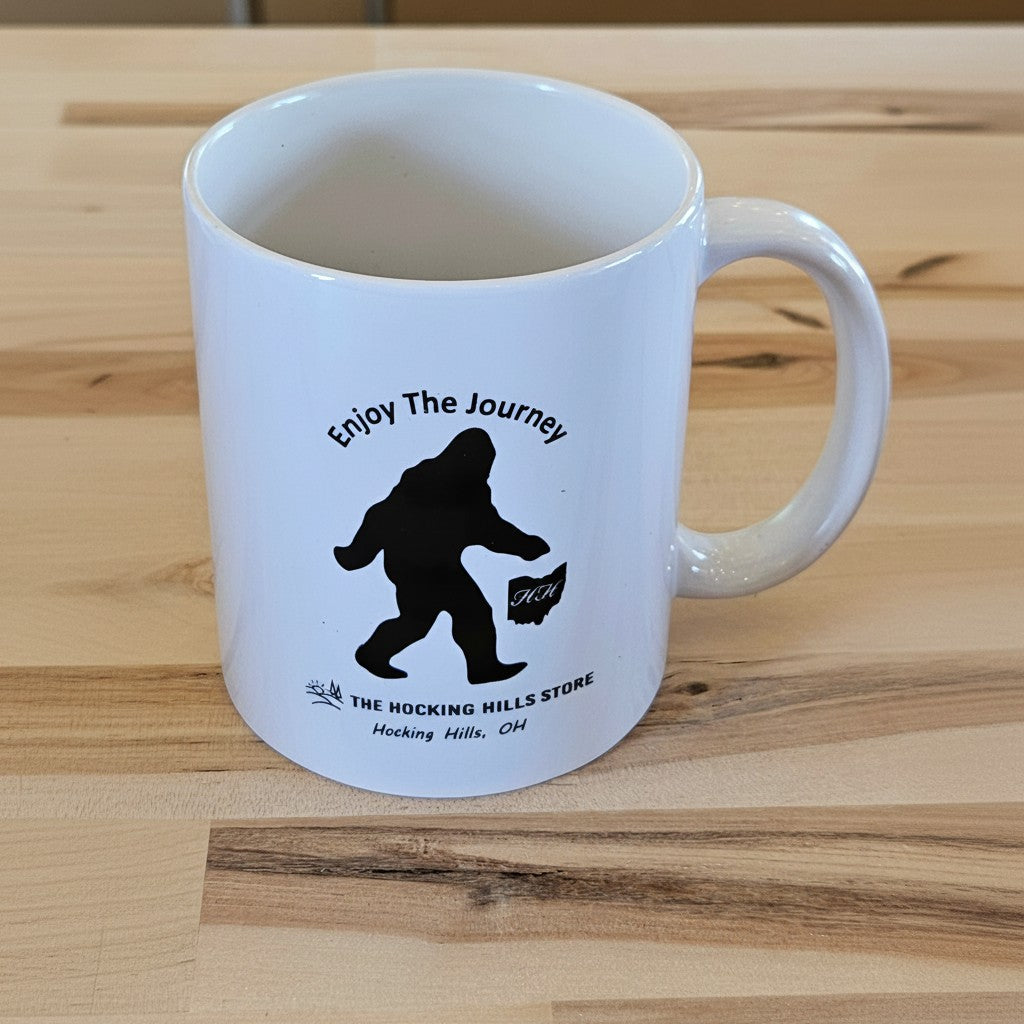 The Hocking Hills Store white ceramic coffee mug with a black Bigfoot silhouette and "Enjoy The Journey" text.