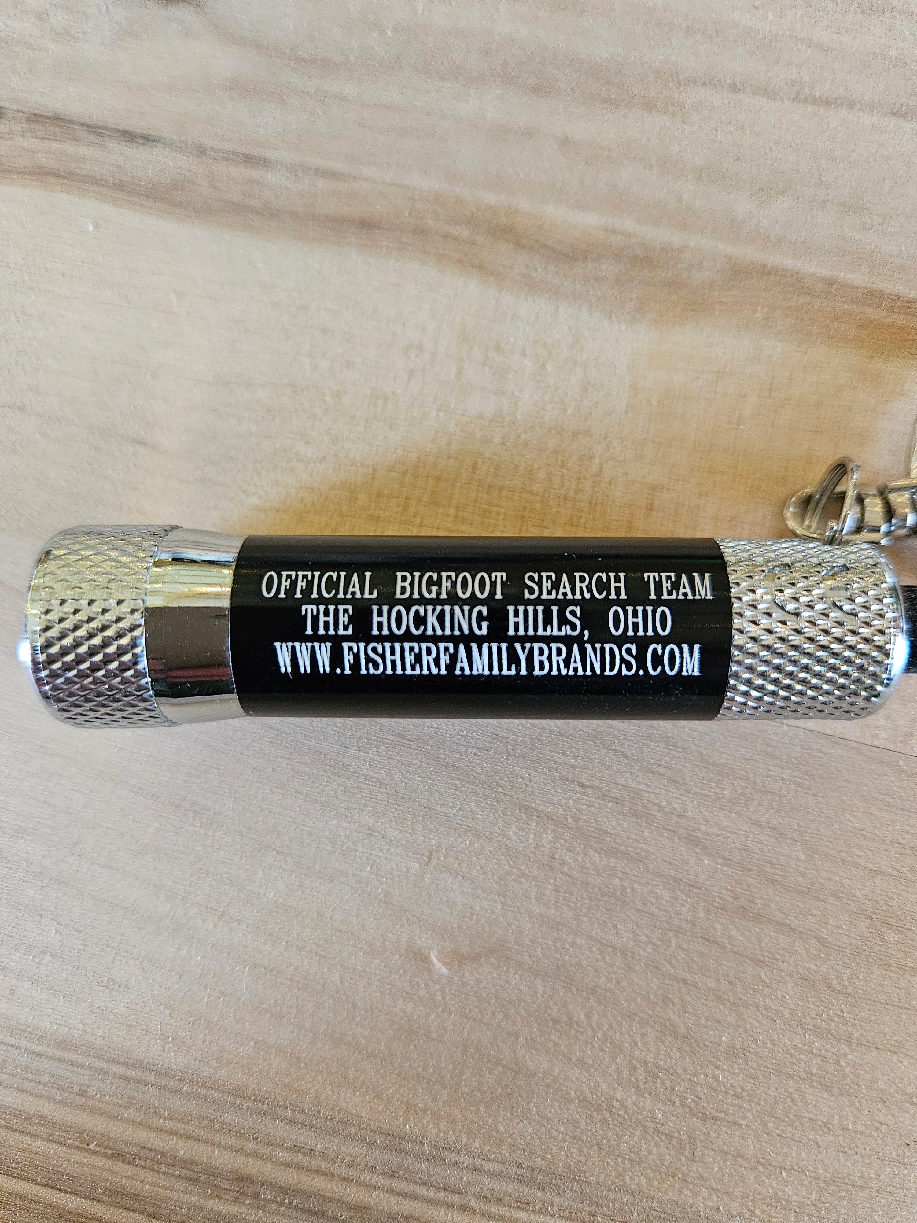 The Hocking Hills Store Bigfoot Search Team mini flashlight keychain with silver textured ends and black body.