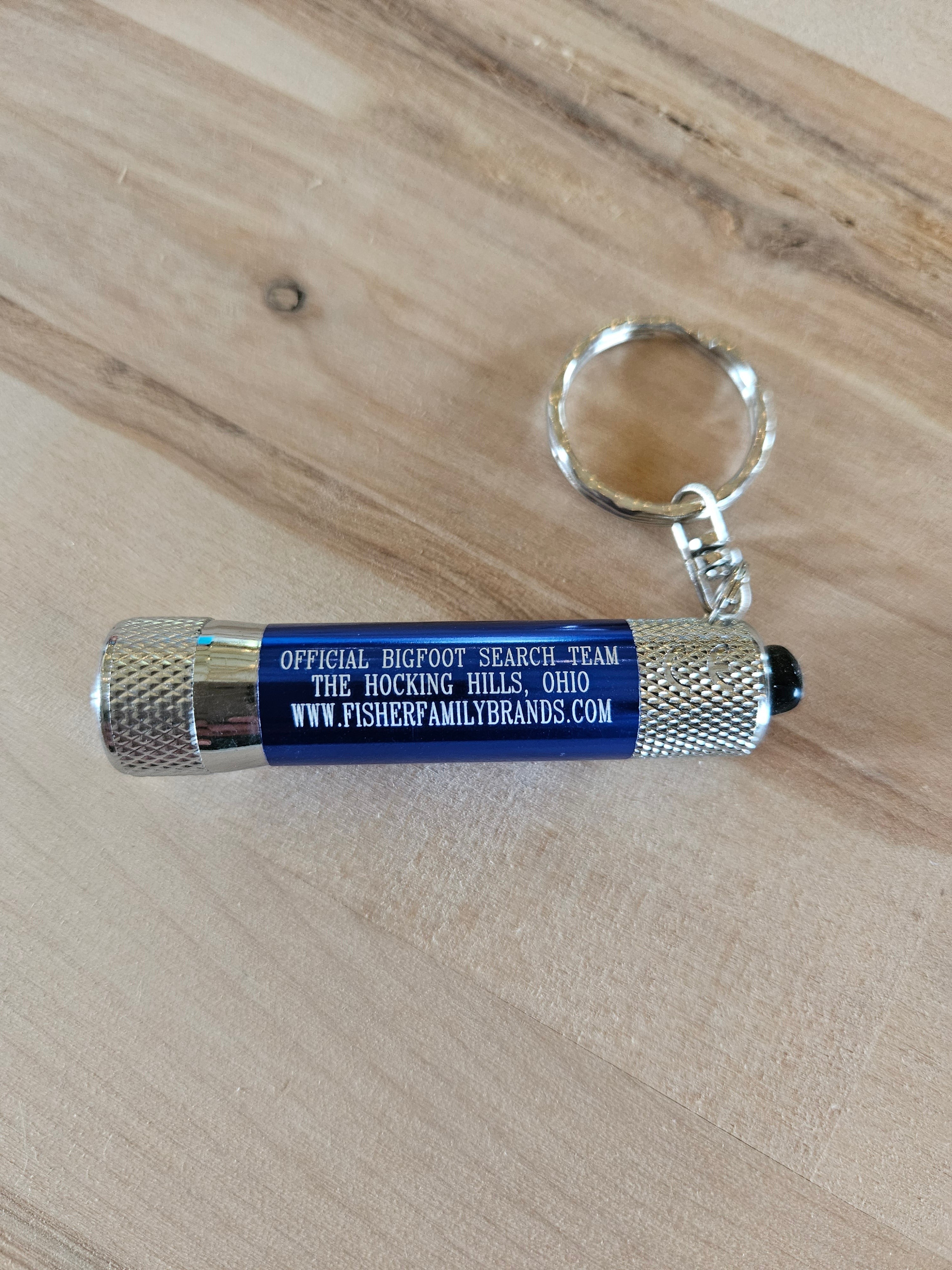 The Hocking Hills Store Bigfoot Search Team mini flashlight keychain with silver metal casing and blue body.