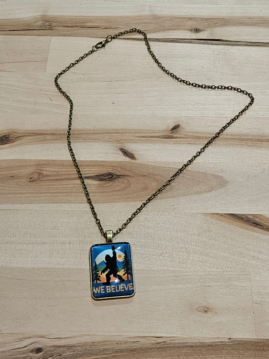 The Hocking Hills Store Bigfoot We Believe pendant necklace with a silhouette of Bigfoot against a sunset landscape.