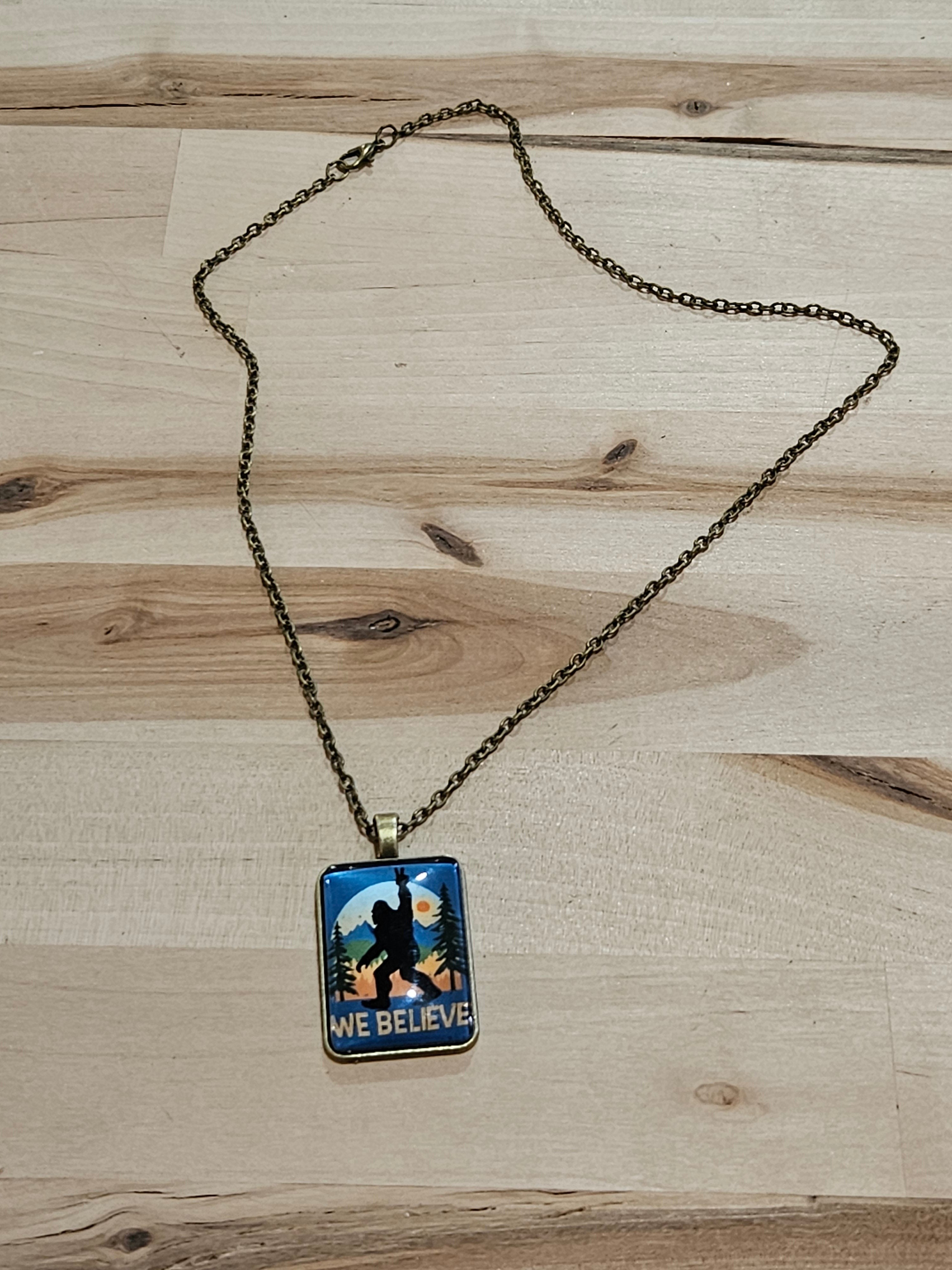 The Hocking Hills Store Bigfoot We Believe pendant necklace with a silhouette of Bigfoot against a sunset landscape.
