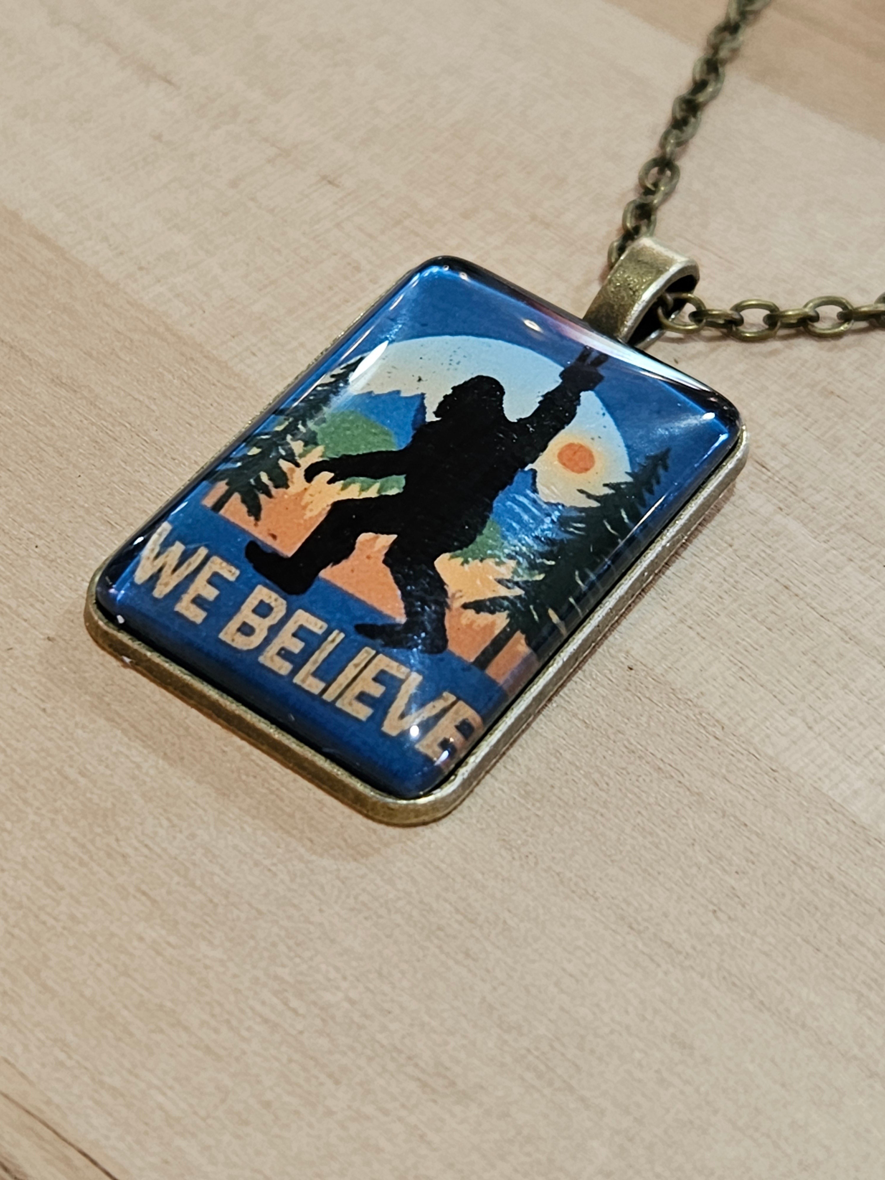 The Hocking Hills Store Bigfoot We Believe pendant with a silhouette of Bigfoot against a sunset and trees. Bronze chain.