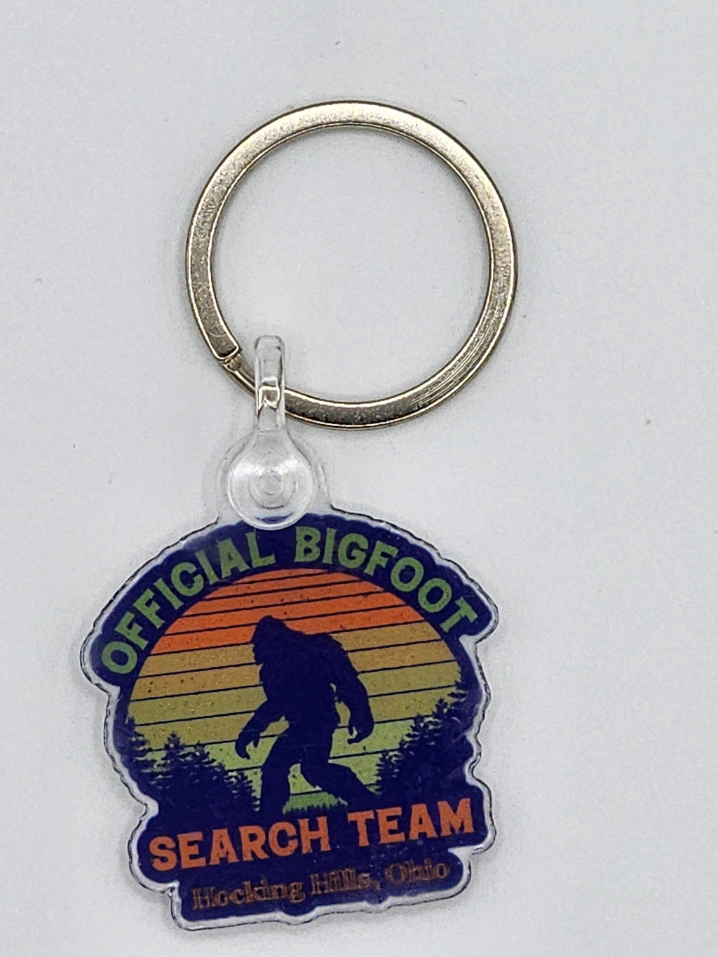 The Hocking Hills Store Official Bigfoot Search Team keychain with a silhouette of Bigfoot against a sunset and forest.