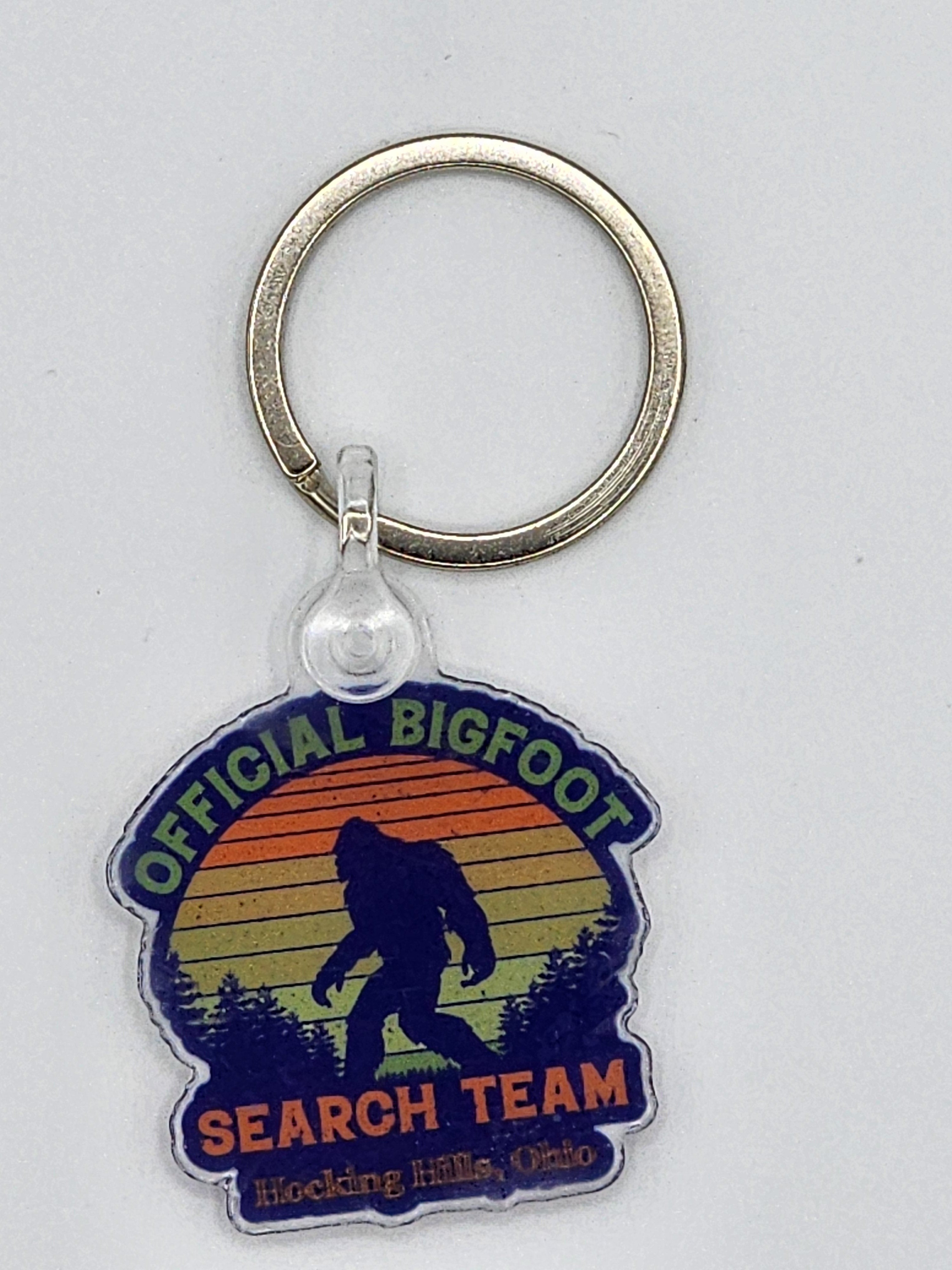 The Hocking Hills Store Official Bigfoot Search Team keychain with a silhouette of Bigfoot against a sunset and forest.