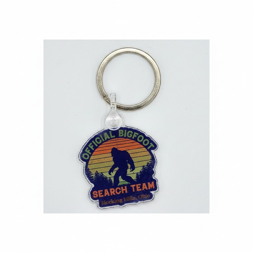 The Hocking Hills Store Official Bigfoot Search Team keychain with a silhouette of Bigfoot against a sunset and forest.