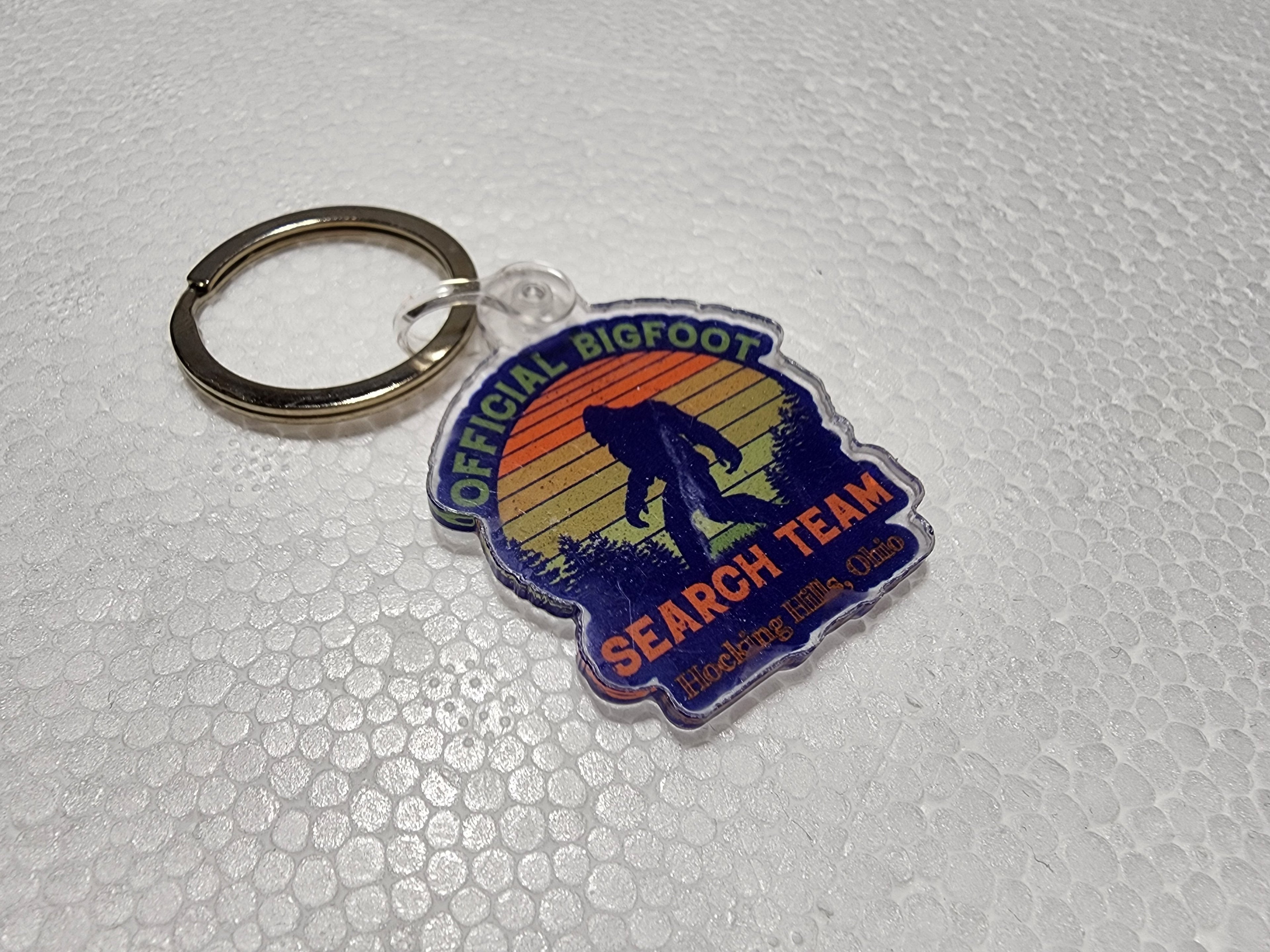 The Hocking Hills Store Official Bigfoot Search Team keychain features a silhouette of Bigfoot against a sunset mountain landscape.