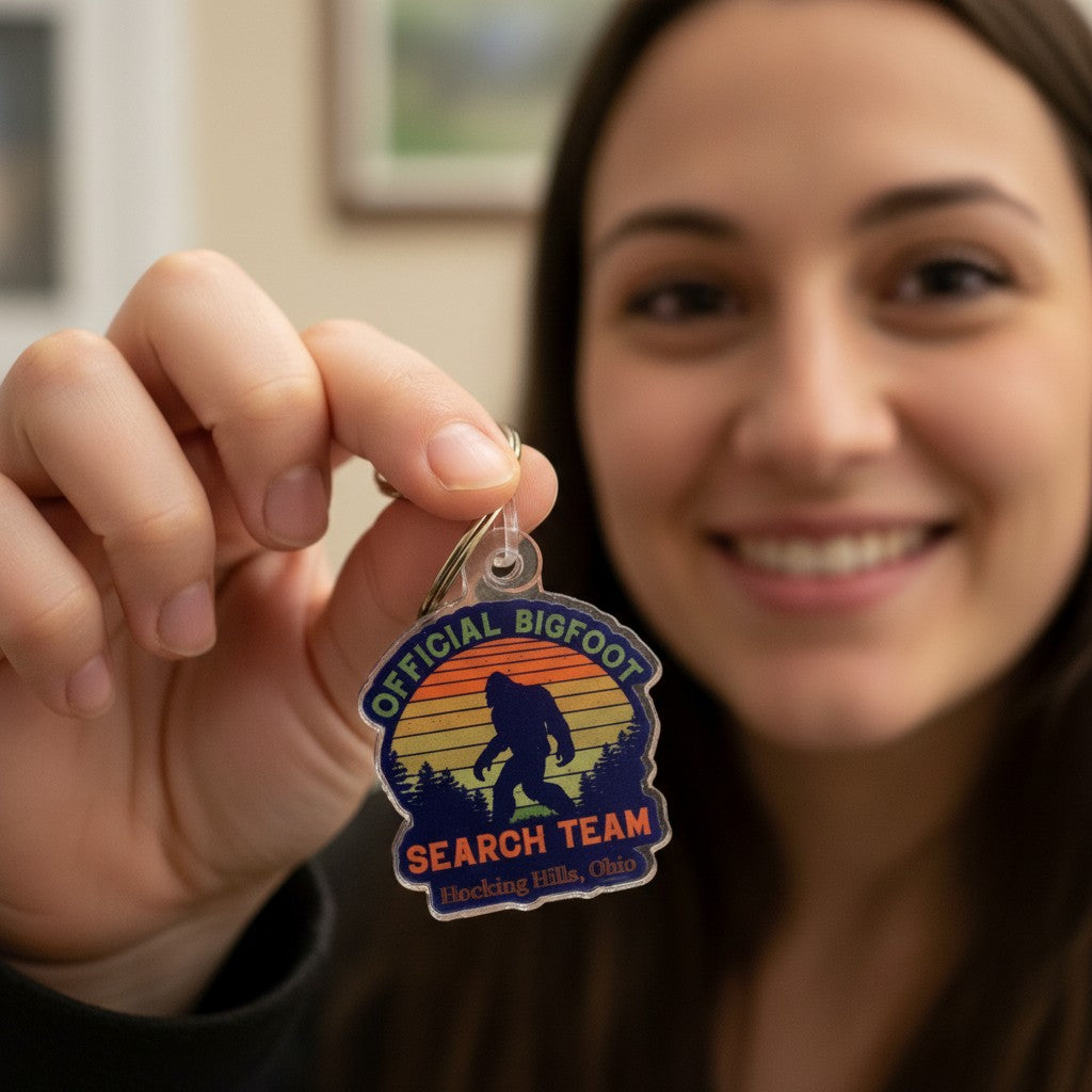 The Hocking Hills Store Official Bigfoot Search Team keychain features a silhouette of Bigfoot against a sunset mountain landscape. Made of clear acrylic.
