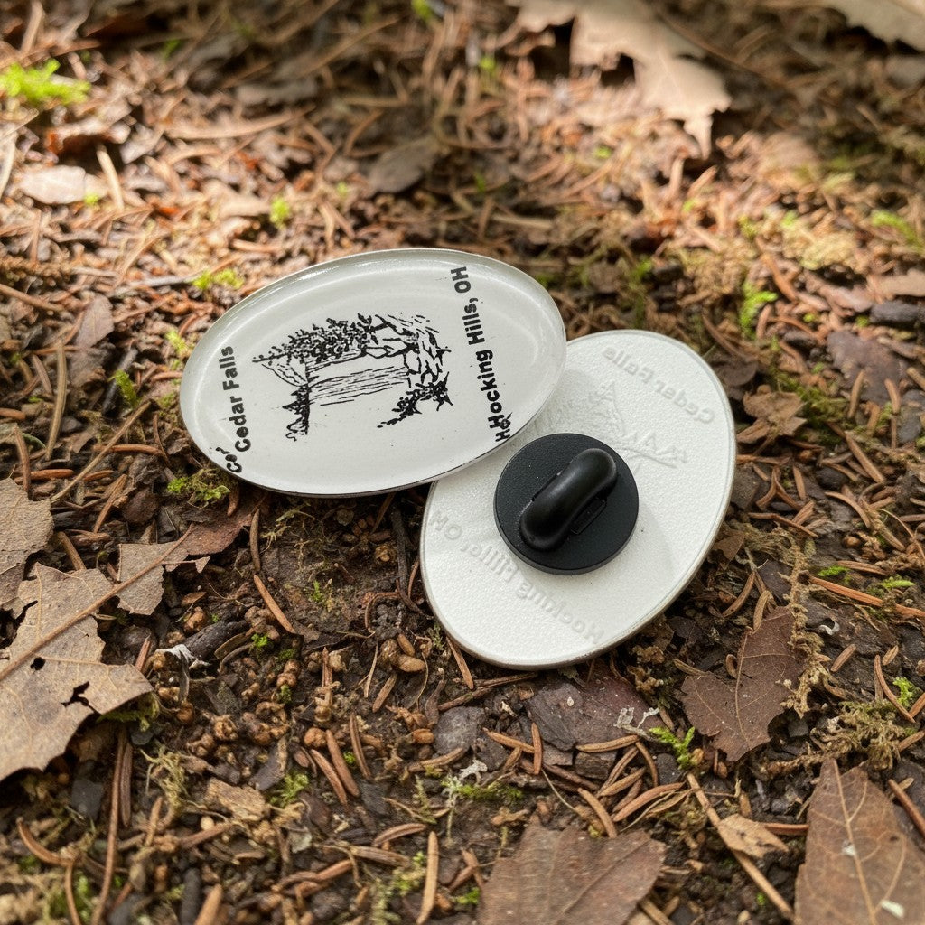 The Hocking Hills Store oval enamel pin featuring a black and white illustration of Cedar Falls waterfall and text.