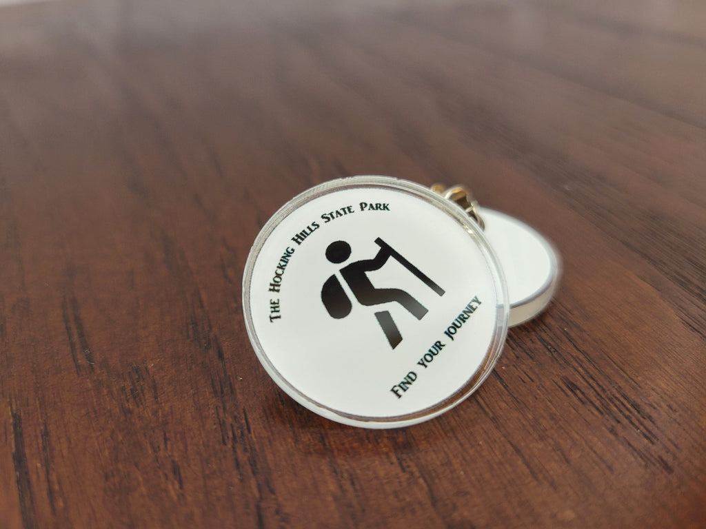 The Hocking Hills Store white round collector pin with hiker graphic and "find your journey" text.