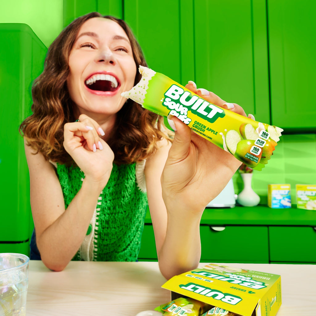 The Hocking Hills Store Green Apple Crush protein bar with a tangy center and crunchy sugar sprinkles, held by a laughing woman.