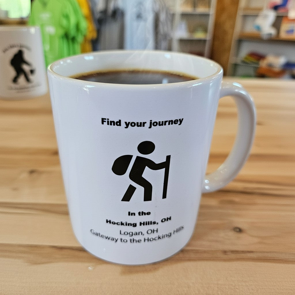 The Hocking Hills Store white ceramic mug with black text and hiker graphic. Features 'Find your journey' and location details.