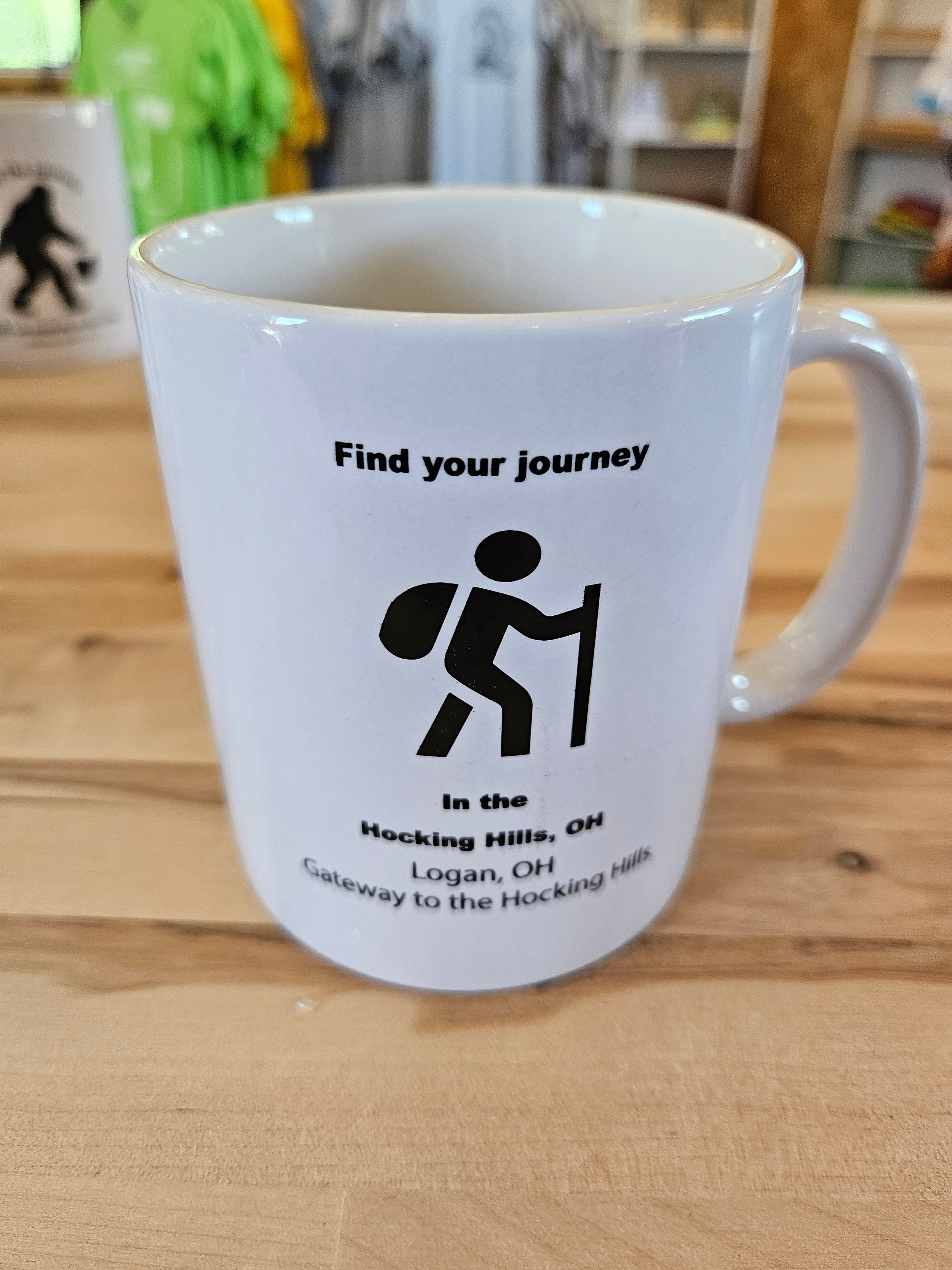 The Hocking Hills Store white ceramic mug with black text and hiker graphic. Text reads "Find your journey" and "In the Hocking Hills, OH Logan, OH Gateway to the Hocking Hills."