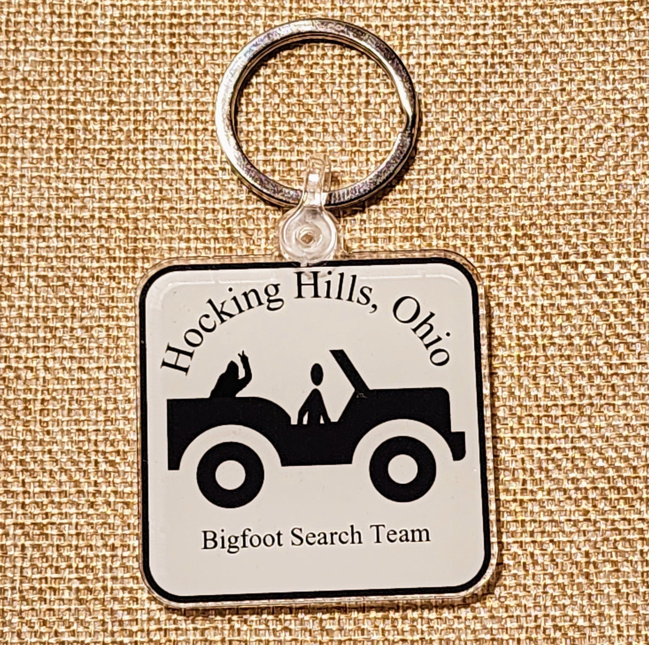 The Hocking Hills Store Jeep Bigfoot Search Team keychain features a black and white graphic of a jeep with two figures inside, text reads "Hocking Hills, Ohio Bigfoot Search Team".