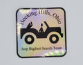 The Hocking Hills Store Jeep Bigfoot Search Team bumper sticker with holographic finish. Features a black Jeep silhouette with two figures and text.