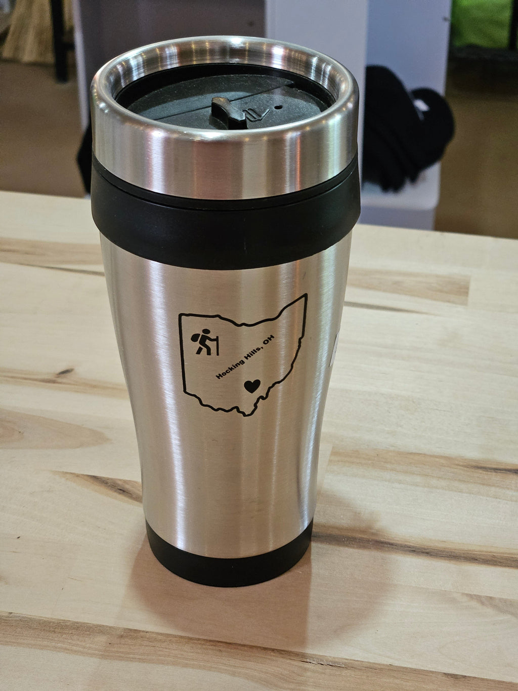 The Hocking Hills Store stainless steel travel mug with black lid and grip. Features Ohio outline with hiking graphic and text.